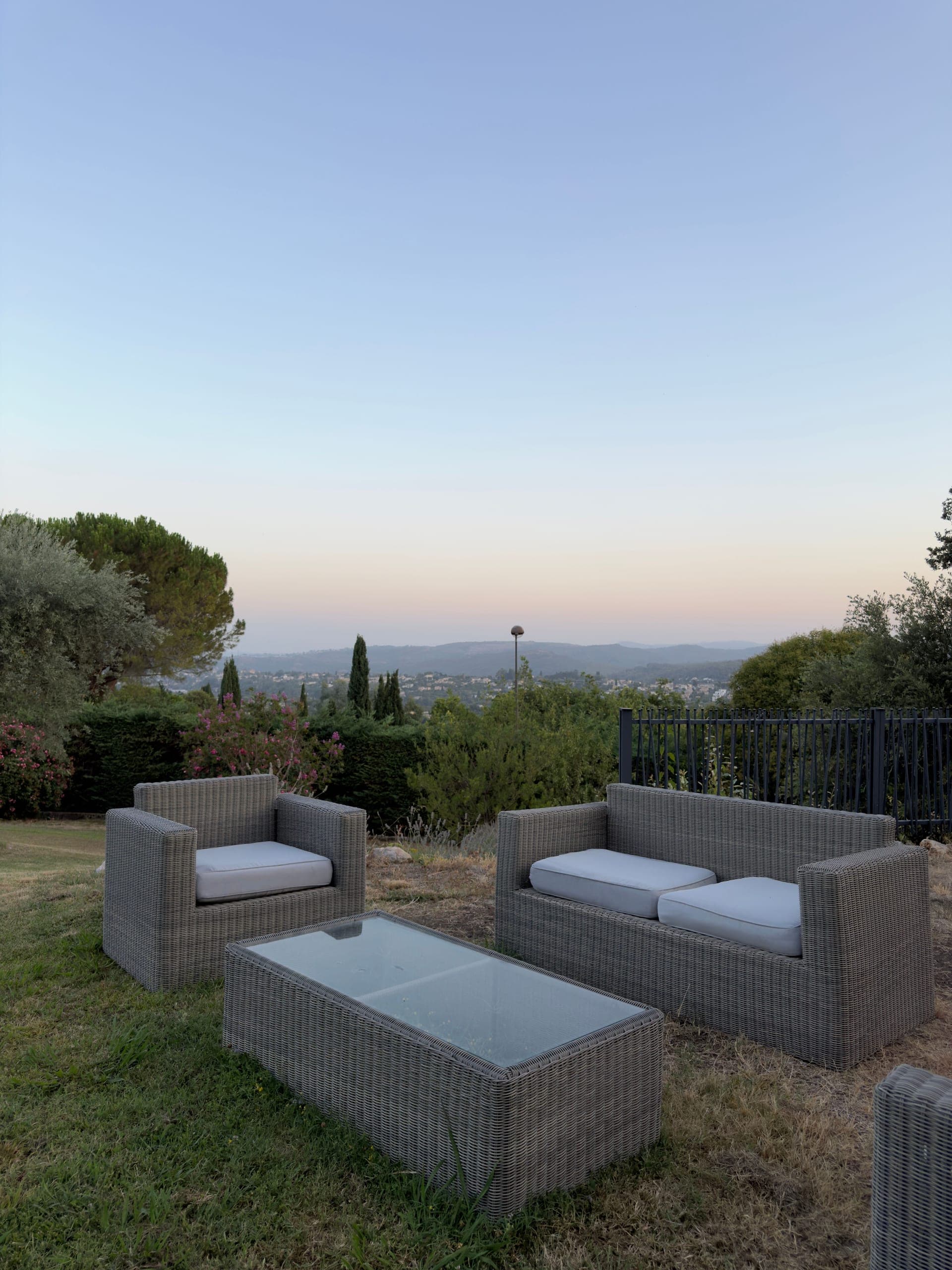 Salon de jardin en résine tressée grise sur pelouse, haie, clôture noire, collines et villages au crépuscule, Saint-Paul-de-Vence
